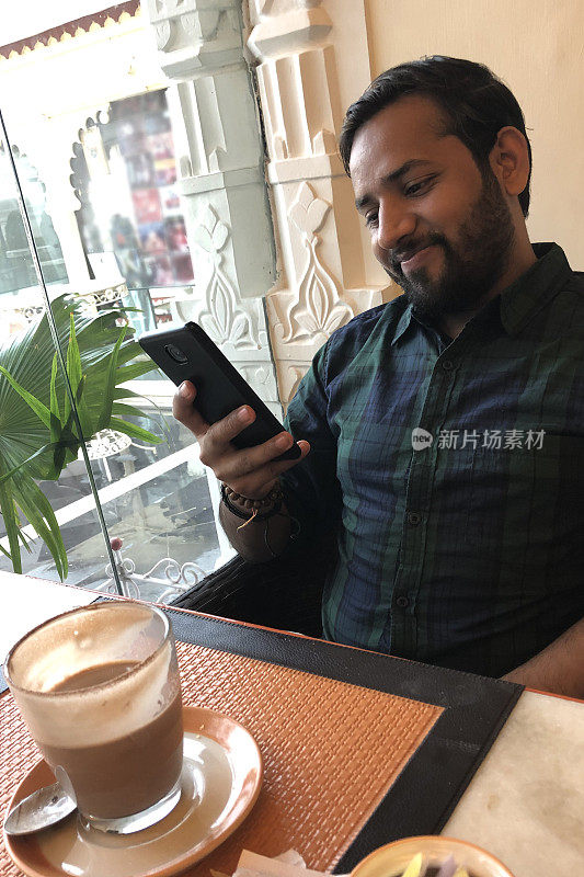 一个快乐的印度人坐在酒店餐厅的早餐桌旁，看着手机屏幕，拿着手机查看社交媒体，互联网新闻，微笑着读短信，喝着咖啡/热巧克力，印度新德里的咖啡馆